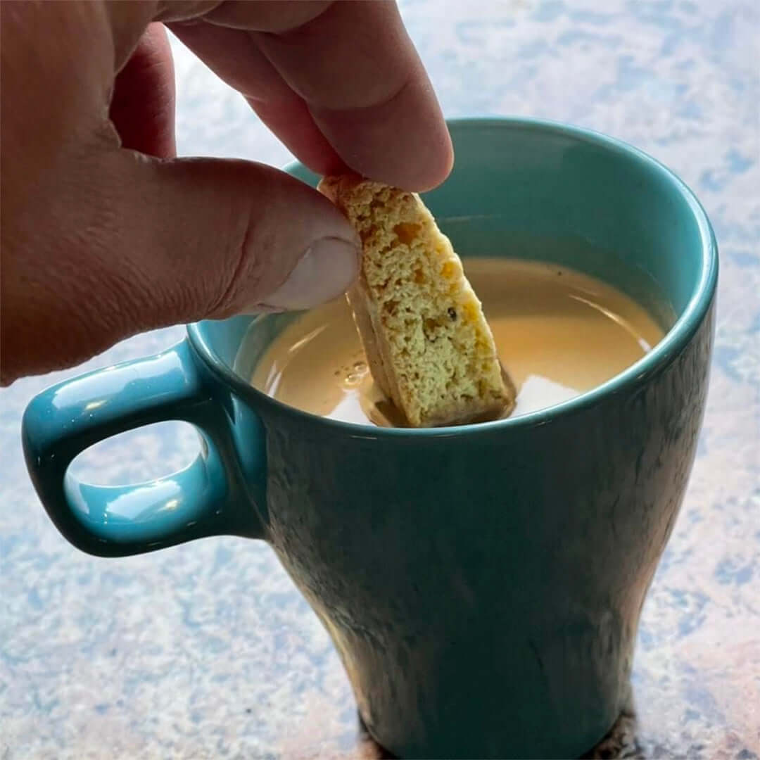A hand dips a piece of True Delicious Whole Almond Biscotti, crafted with premium California almonds from a classic Tuscan recipe, into a teal mug of coffee or tea on a tabletop.