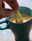 A hand dips a piece of True Delicious Whole Almond Biscotti, crafted with premium California almonds from a classic Tuscan recipe, into a teal mug of coffee or tea on a tabletop.
