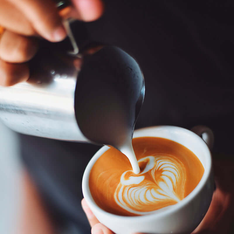 A hand pours steamed milk into a cup made with True Delicious Coffee Beans for Espresso 1lb bag, creating heart-shaped latte art on the coffee’s surface.