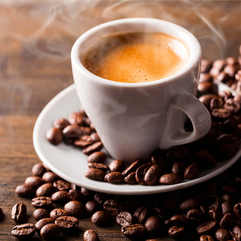 A steaming cup of coffee in a white cup and saucer sits on a wooden table, surrounded by scattered True Delicious Coffee Beans for Espresso 1lb bag.