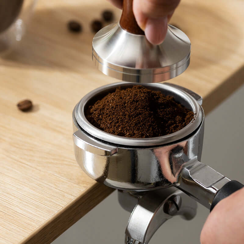 A hand presses a tamper onto ground coffee in a portafilter, preparing True Delicious Coffee Beans for Espresso (1lb bag) on a light wooden surface. Premium beans are scattered nearby.