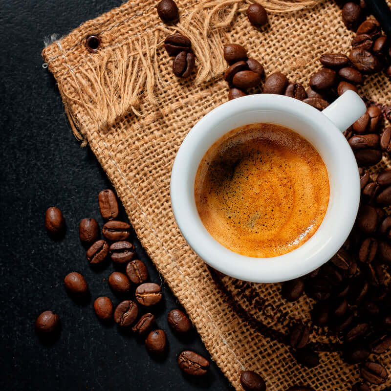 A white cup of espresso sits on burlap, surrounded by True Delicious Coffee Beans for Espresso from a 1lb bag, with scattered premium beans on a dark surface.