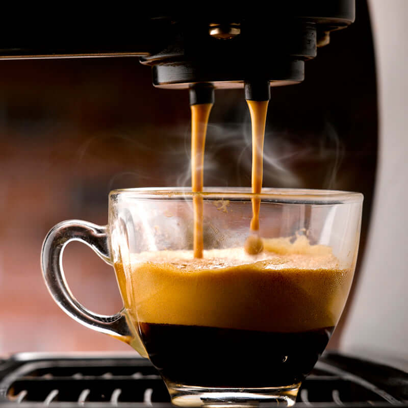 A close-up of an espresso machine pouring hot coffee made with True Delicious Coffee Beans for Espresso 1lb bag into a clear glass cup, highlighting the rich crema and steam rising from freshly brewed premium beans.