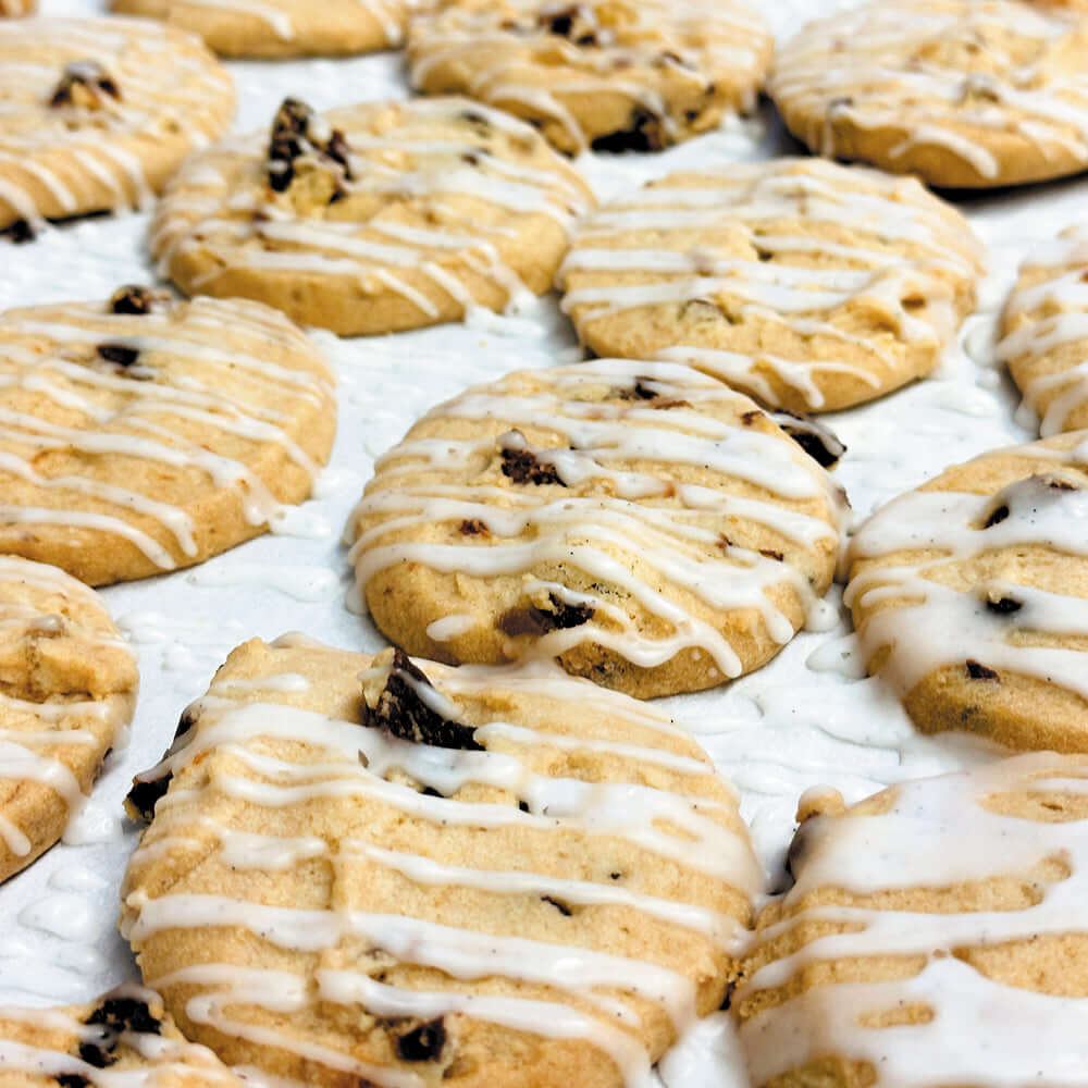 Close-up of True Delicious Apricot-Ginger Cookies with Vanilla Bean Glaze—round shortbread cookies featuring chocolate chunks and crystallized ginger, drizzled with white icing on a white textured surface.