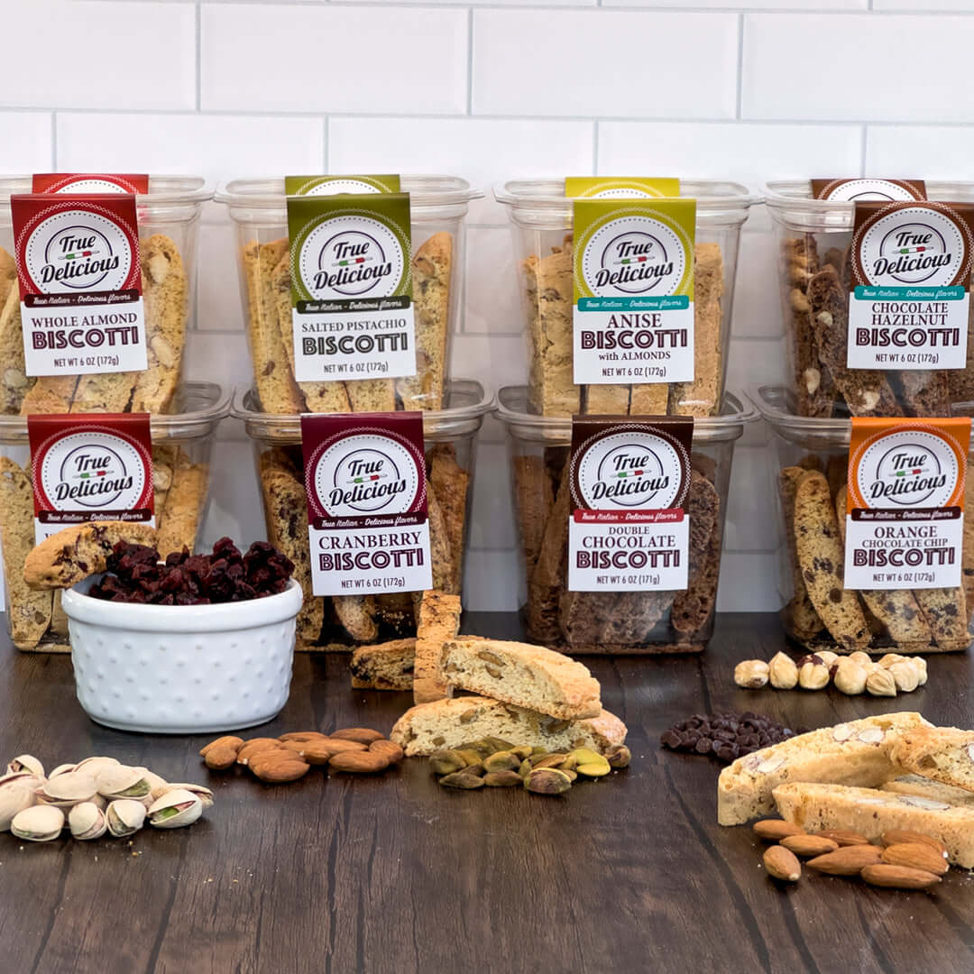 Eight packs of True Delicious All Flavors Biscotti 8-Pack are arranged on a wooden surface with scattered biscotti pieces, nuts, and cranberries. Handcrafted in Petaluma with non-GMO ingredients, theyre set against a white tiled wall.