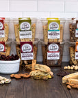 Eight packs of True Delicious All Flavors Biscotti 8-Pack are arranged on a wooden surface with scattered biscotti pieces, nuts, and cranberries. Handcrafted in Petaluma with non-GMO ingredients, theyre set against a white tiled wall.