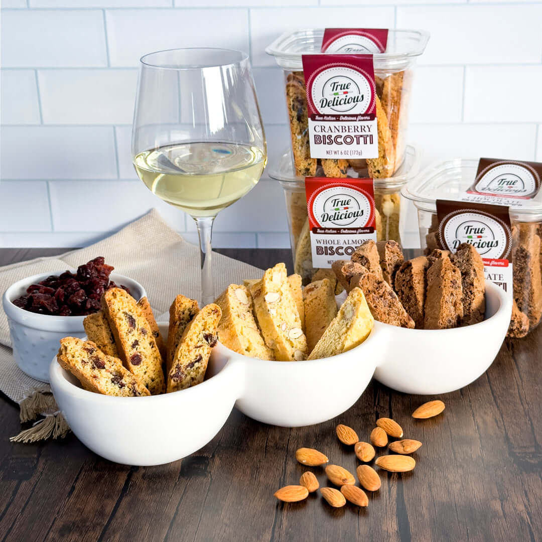 A glass of white wine, three bowls with assorted biscotti from the True Delicious All Flavors Biscotti 8-Pack, plus dried cranberries and whole almonds, are arranged on a wooden surface against a white tiled background.