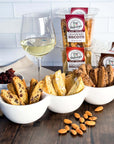 A glass of white wine, three bowls with assorted biscotti from the True Delicious All Flavors Biscotti 8-Pack, plus dried cranberries and whole almonds, are arranged on a wooden surface against a white tiled background.