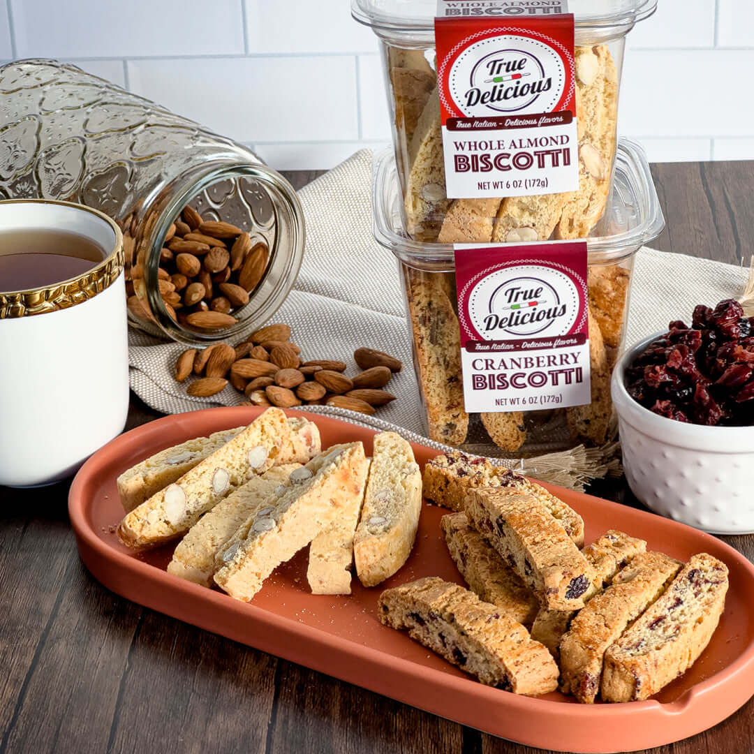 A plate of handcrafted True Delicious Cranberry Biscotti sits beside tea, almonds, dried cranberries, and containers labeled “True Delicious Cranberry Biscotti,” made with natural ingredients.
