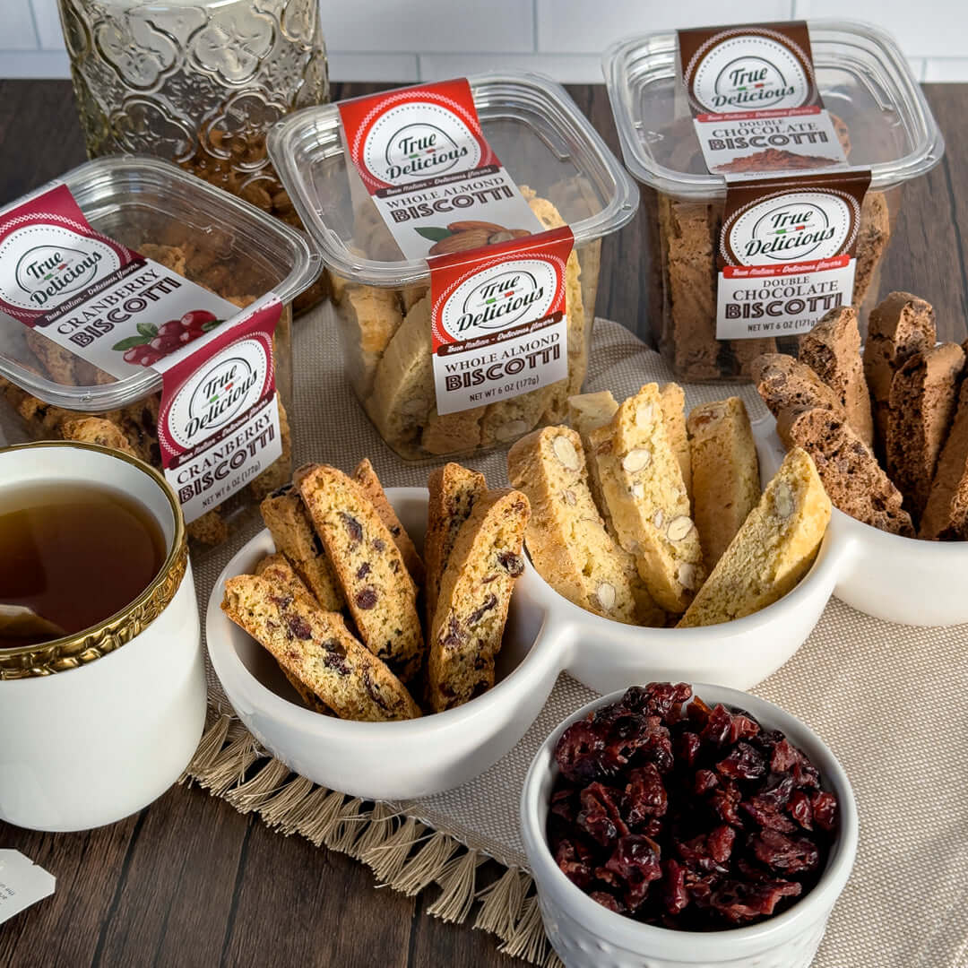 True Delicious All Flavors Biscotti 8-Pack, made with non-GMO ingredients, is displayed in containers and bowls beside dried cranberries and a cup of tea on a wooden surface with a beige napkin.
