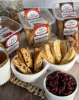 True Delicious All Flavors Biscotti 8-Pack, made with non-GMO ingredients, is displayed in containers and bowls beside dried cranberries and a cup of tea on a wooden surface with a beige napkin.