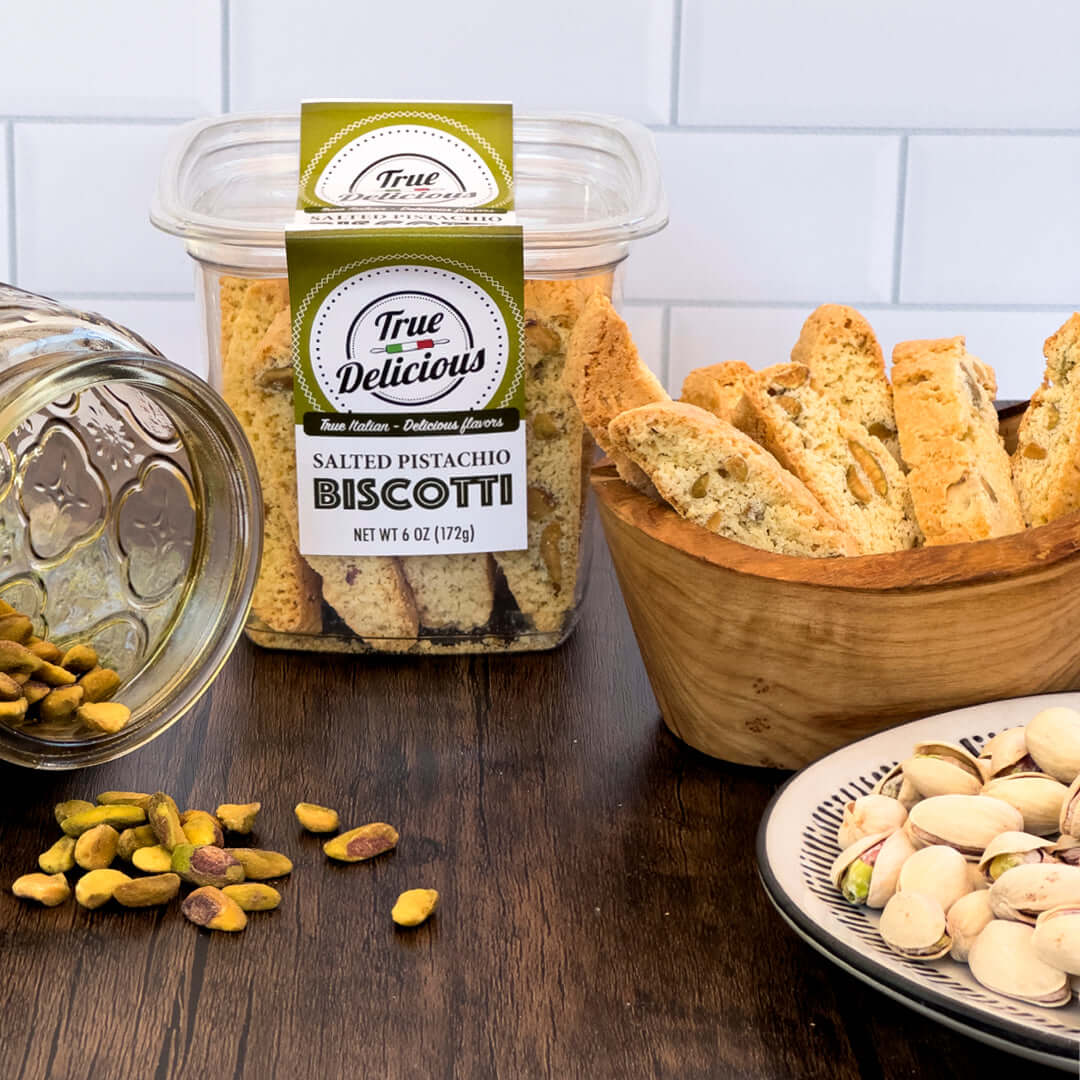 On a wooden table against a white tiled background, True Delicious Salted Pistachio Biscotti sits in a labeled tub alongside artisanal biscotti in a bowl, a jar of spilled pistachios, and a plate of shelled pistachios.