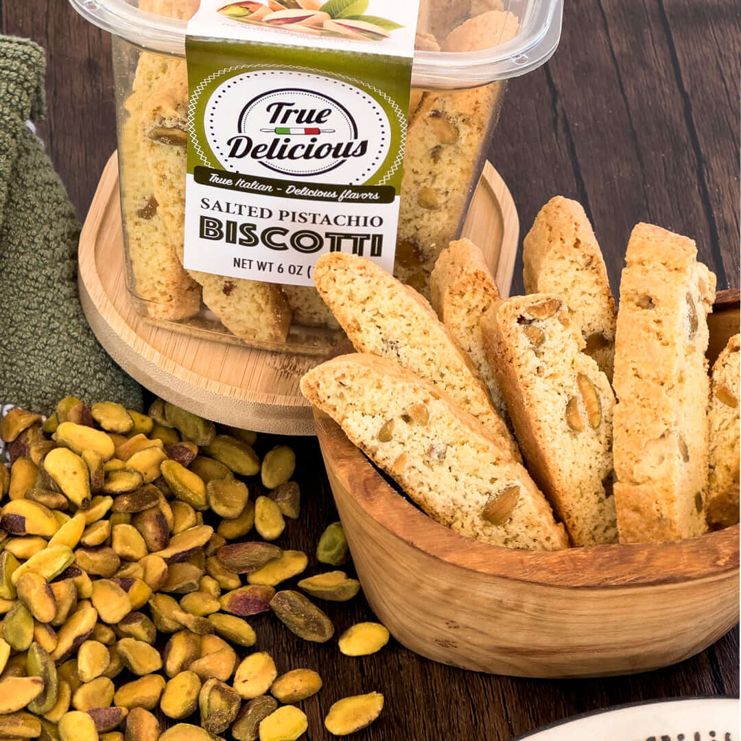 A container of True Delicious Salted Pistachio Biscotti sits on a wooden surface next to a wooden bowl filled with biscotti and scattered shelled pistachios.