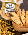 A container of True Delicious Salted Pistachio Biscotti sits on a wooden surface next to a wooden bowl filled with biscotti and scattered shelled pistachios.