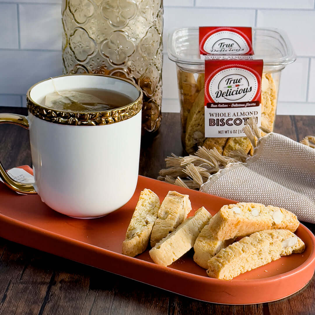 A gold-rimmed cup of coffee sits on an orange tray beside several pieces of True Delicious Whole Almond Biscotti, made with premium California almonds. In the background is a container labeled True Delicious on a wooden table.