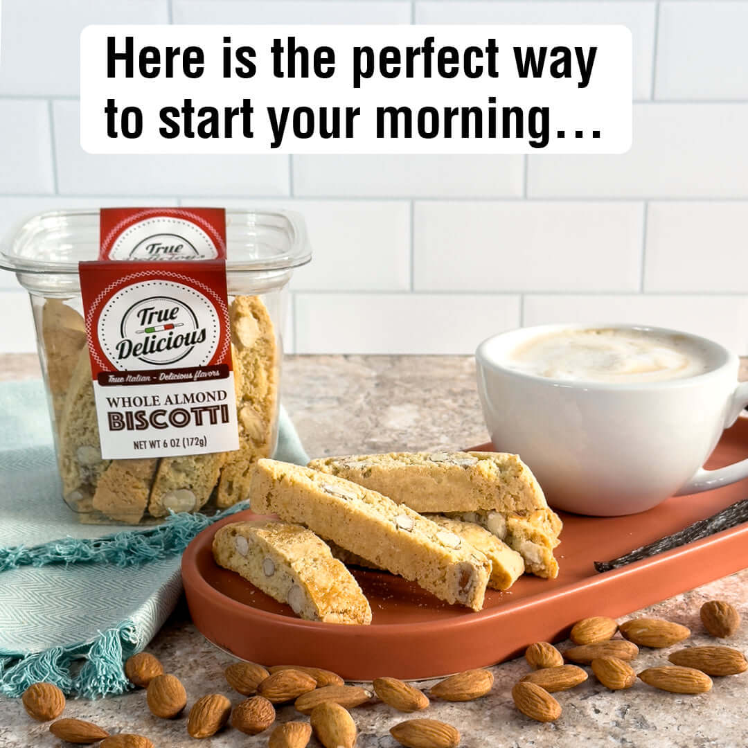 A plate with a cappuccino and three True Delicious Whole Almond Biscotti—crafted from a Tuscan recipe with California almonds—rests on a table with scattered almonds. The product container sits behind, ready for your perfect morning start.