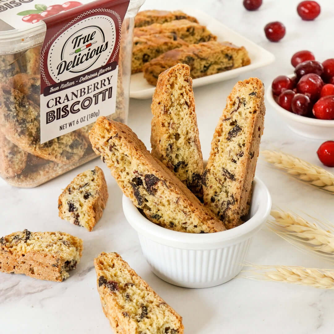 A small white bowl holds several pieces of True Delicious Cranberry Biscotti. More biscotti and a clear container labeled True Delicious Cranberry Biscotti sit in the background, with dried cranberries and wheat stalks nearby.