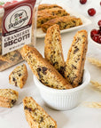 A small white bowl holds several pieces of True Delicious Cranberry Biscotti. More biscotti and a clear container labeled True Delicious Cranberry Biscotti sit in the background, with dried cranberries and wheat stalks nearby.