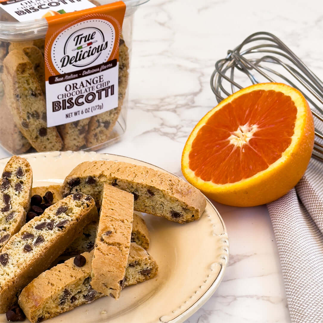 A plate with orange chocolate chip biscotti, chocolate chips, a sliced orange half, and a True Delicious Bundle of the Month container—made with locally sourced ingredients for the finest Biscotti and Cookies experience.