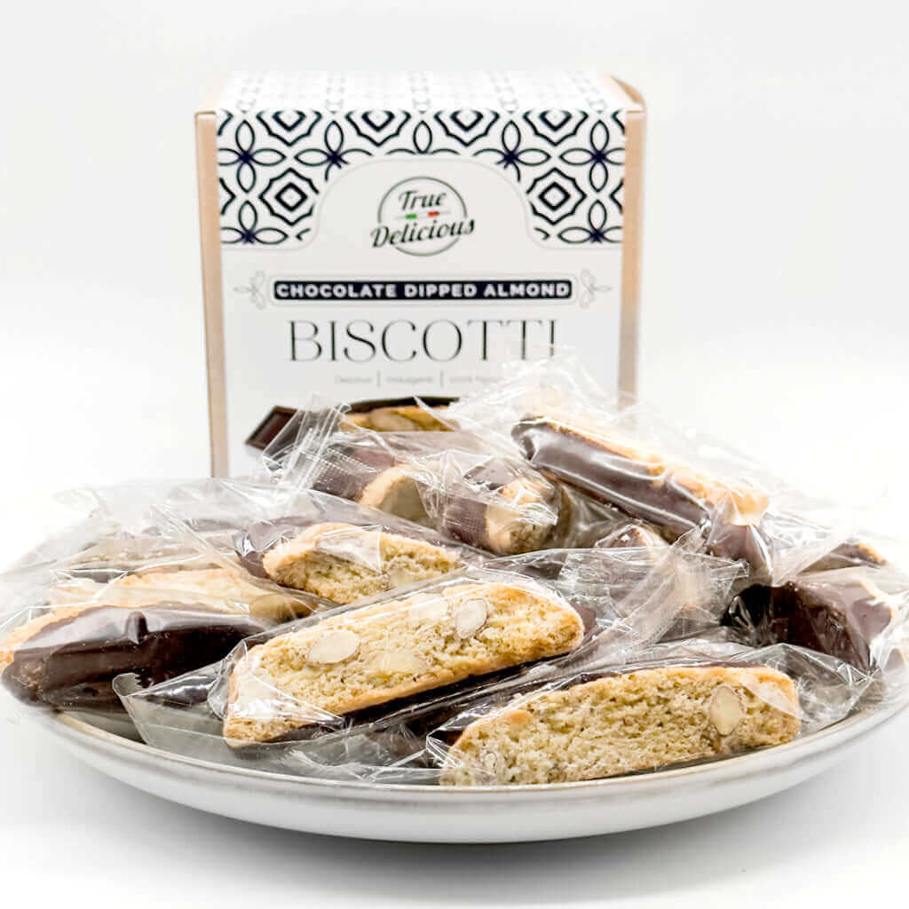 A plate holds individually wrapped 2x Chocolate Dipped Almond Biscotti from True Delicious, displayed in front of a True Delicious box. The artisanal biscotti are clearly visible through the transparent wrappers.