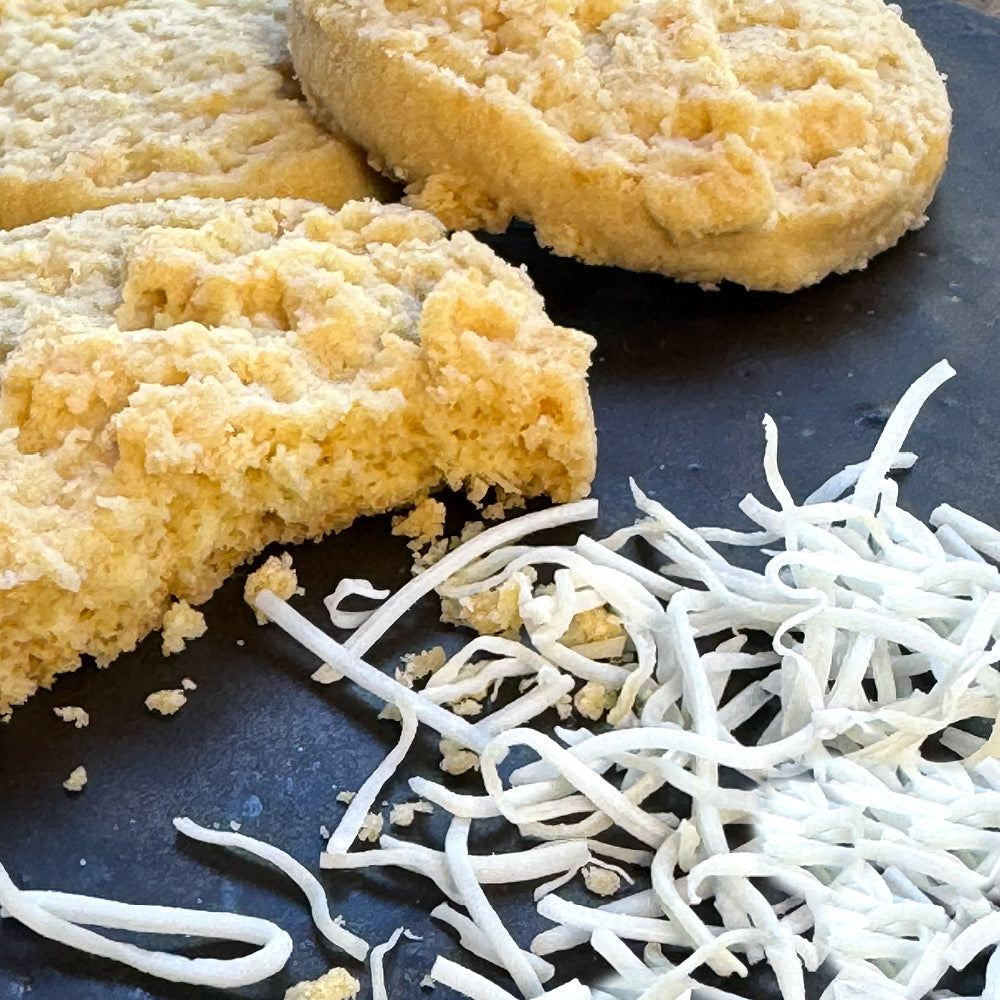 Three golden Coconut Shortbread Cookies by True Delicious Biscotti rest on a dark surface, one partially broken, with shredded coconut in the foreground for added texture and flavor.
