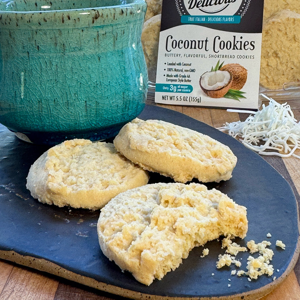 Three coconut cookies, one partially eaten, rest on a dark plate near a turquoise mug. Behind them, a box labeled True Delicious Biscotti Coconut Shortbread Cookies suggests these artisanal treats are topped with shredded coconut.