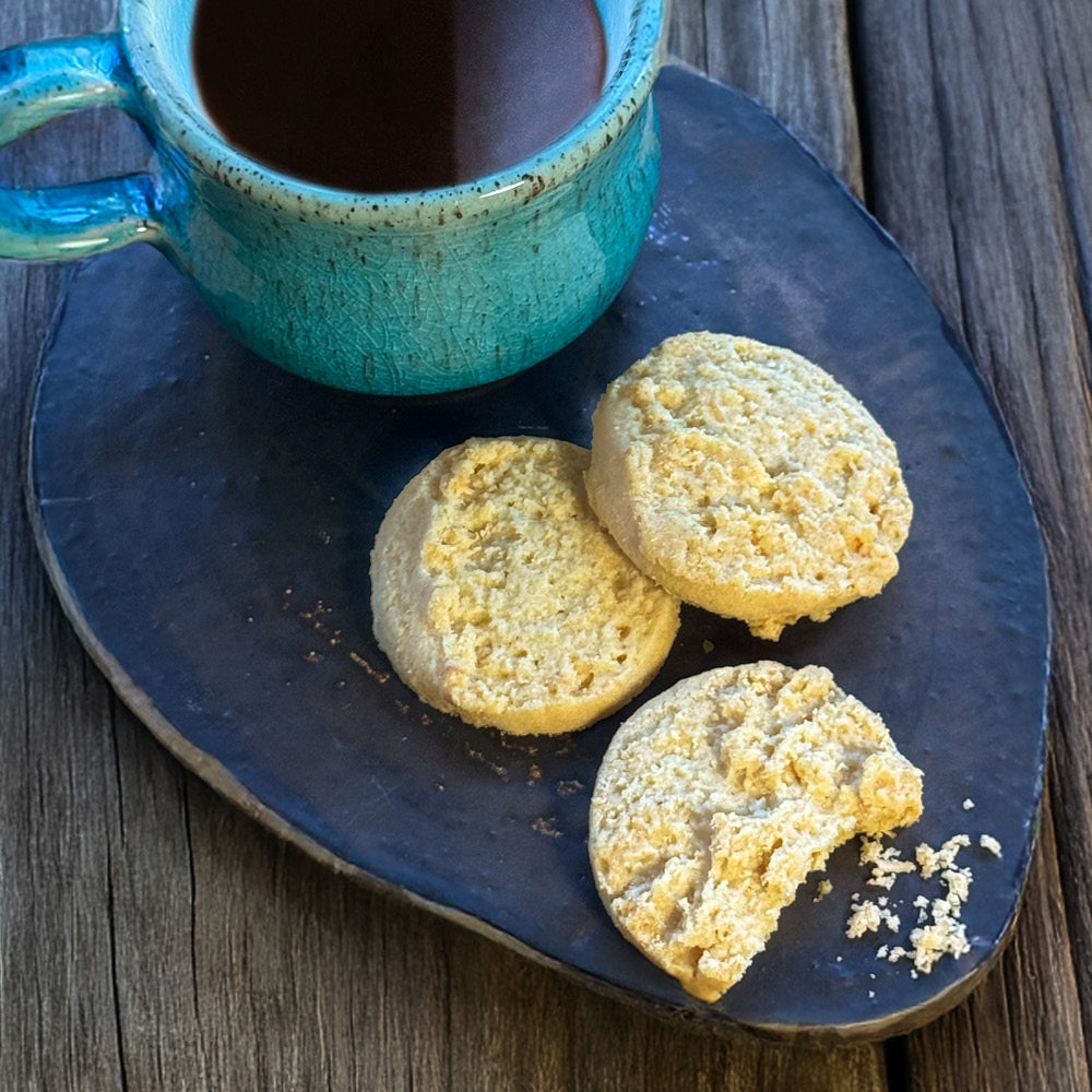 A turquoise coffee cup rests on a dark plate with three True Delicious Biscotti Coconut Shortbread Cookies, one partially eaten, on a rustic wooden surface.