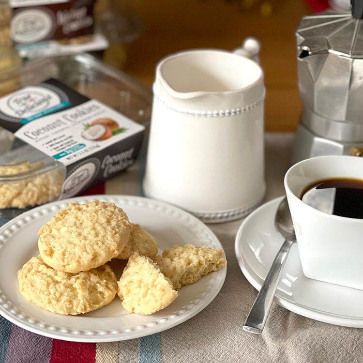 A plate of True Delicious Biscotti’s Coconut Shortbread Cookies sits beside a cup of black coffee, a silver moka pot, and a white creamer on a striped tablecloth.