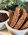 A white bowl holds several True Delicious Double Chocolate Biscotti, standing upright beside a bowl of chocolate chips, with a green leafy plant in the background.