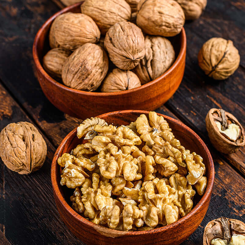 Two wooden bowls—one with whole walnuts, the other with walnut halves—rest on a dark wood surface. Scattered nuts add rustic charm, evoking the nutty sweetness of True Delicious Biscotti Walnut Shortbread Cookies.