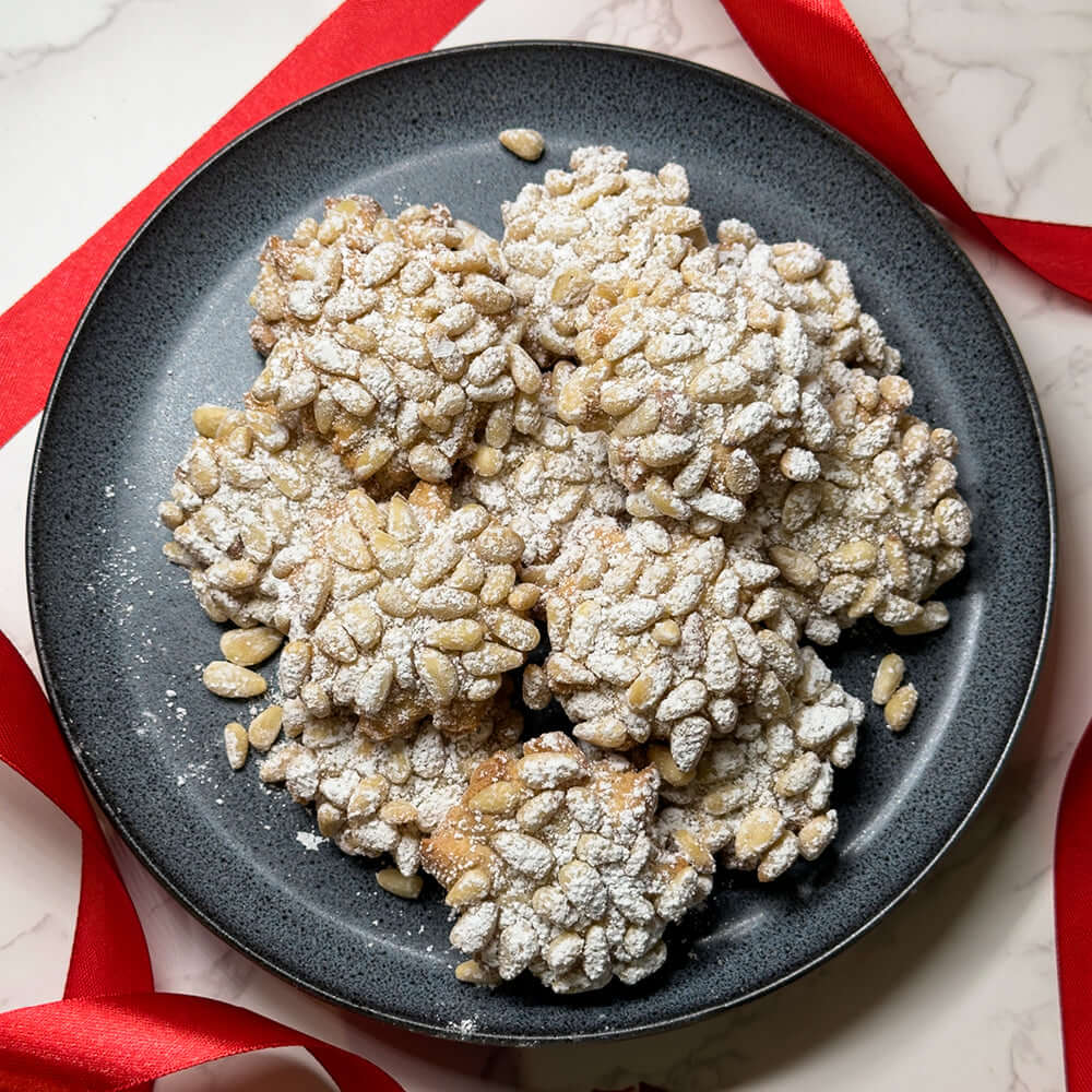 A dark plate displays several Papas Pignoli Cookies by True Delicious, dusted with powdered sugar and accented by red ribbons on a marble surface.