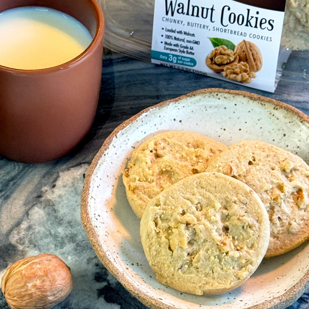 A ceramic plate with three Walnut Shortbread Cookies by True Delicious Biscotti rests on a marbled surface near a whole walnut, a cup of milk, and a package labeled Walnut Cookies, showcasing these delicious artisanal treats.