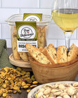 The True Delicious Bundle of the Month features biscotti and cookies in a wooden bowl, a glass of white wine, a Salted Pistachio Biscotti container with locally sourced ingredients, and scattered pistachios on a wooden table with a green towel.