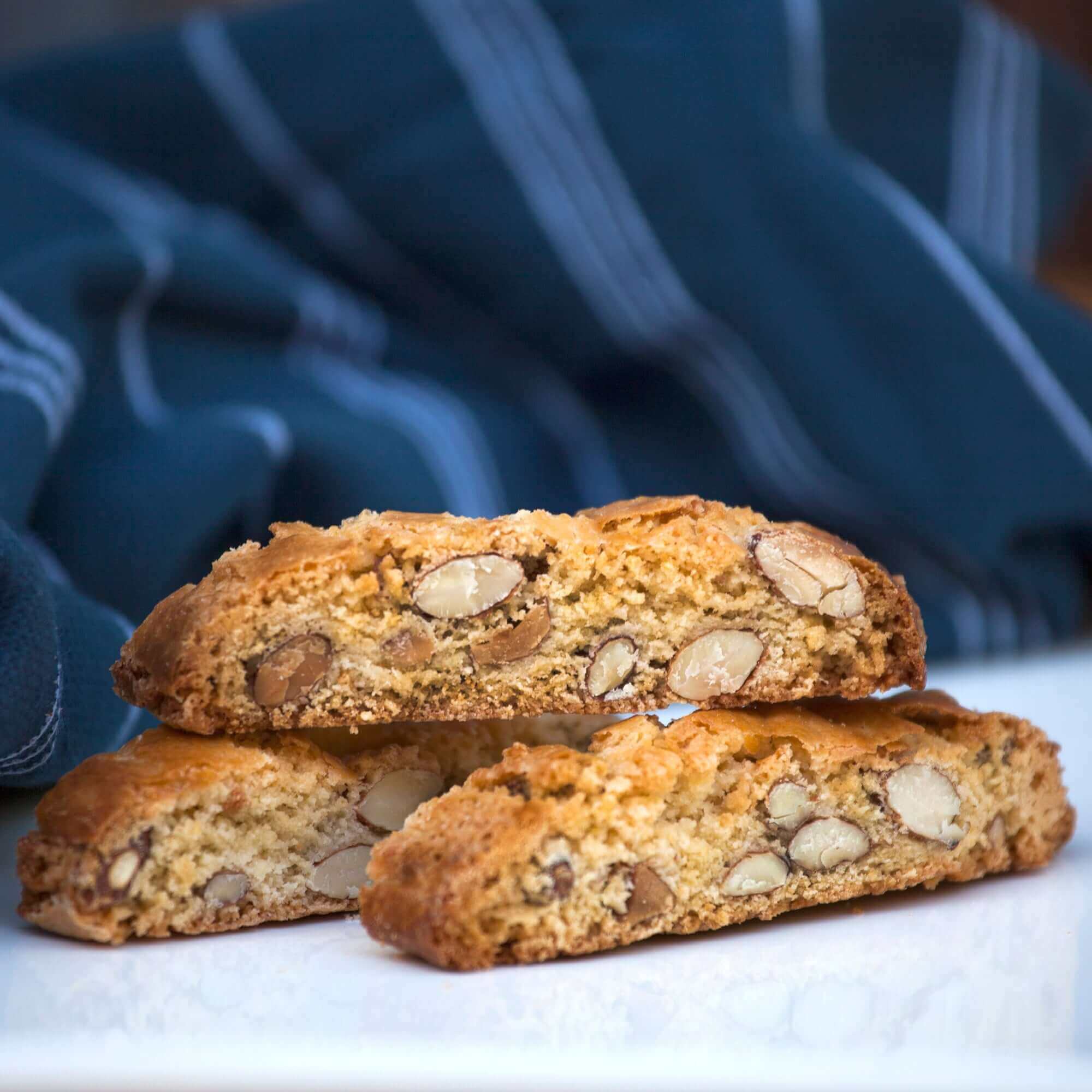 Three True Delicious Whole Almond Biscotti are stacked on a white surface with a blue and white striped cloth in the background. These golden brown treats feature premium California almonds and a classic Tuscan recipe.