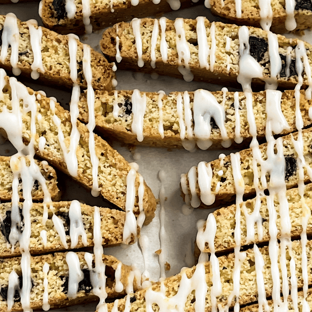 Close-up of True Delicious Lemon Blueberry Biscotti with Glaze, featuring visible nuts and dried fruit, arranged on parchment paper and drizzled with a sweet lemon glaze.