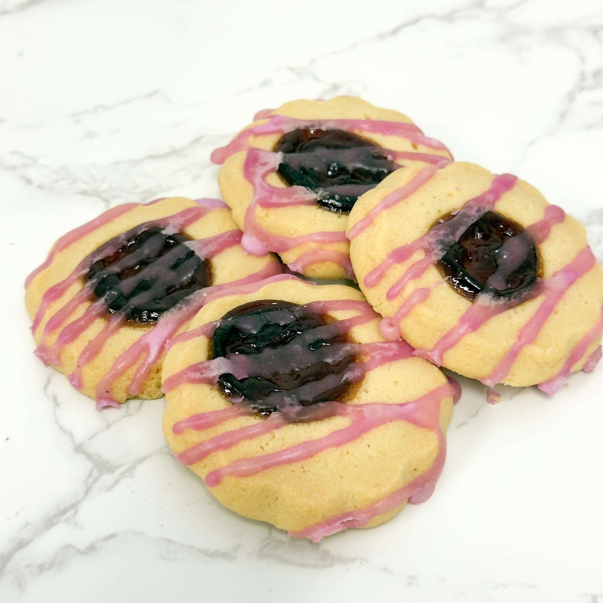 Four buttery True Delicious Jam Cookies - Raspberry, filled with dark raspberry jam and topped with pink icing drizzle, arranged on a white marble surface.