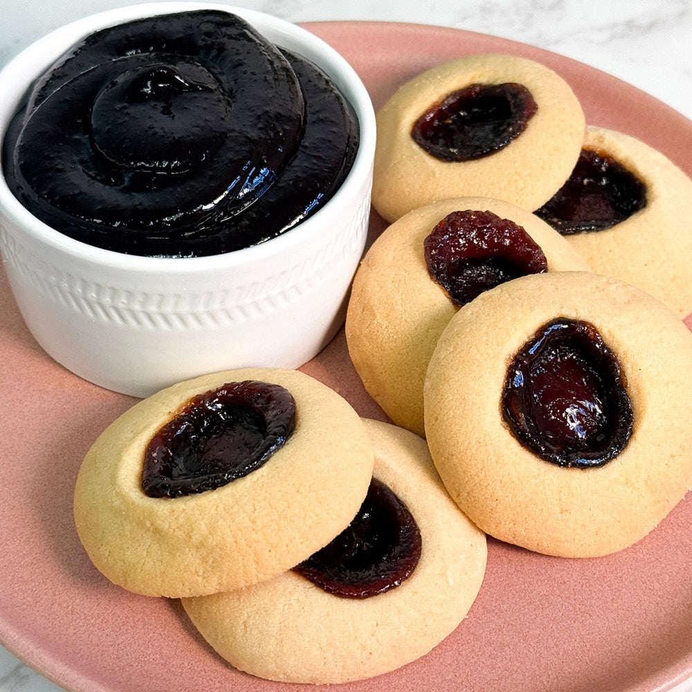 A plate of True Delicious Mixed Berries Jam Cookies with a small white bowl of jam is served on a pink plate.