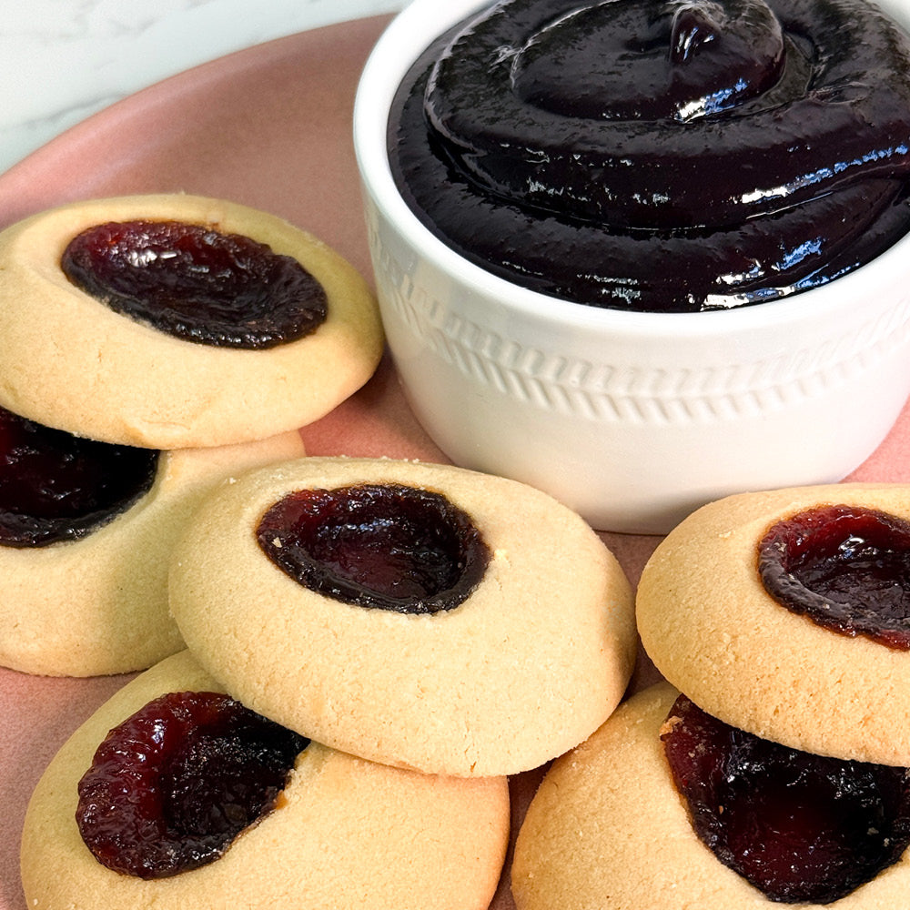 A plate of True Delicious Mixed Berries Jam Cookies, with buttery shortbread and jam centers, sits beside a white bowl brimming with thick fruit spread.