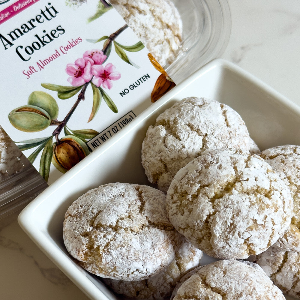 A white bowl of powdered sugar-coated Italian cookies sits next to a True Delicious Amaretti container labeled "Soft Almond Cookies, No Gluten," with packaging featuring a pink flower and almonds.