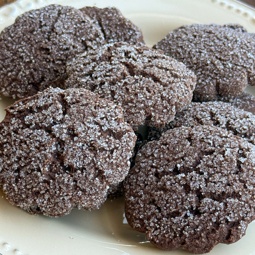 A close-up of True Delicious Cocoa Blooms Cookies, made with natural cocoa and topped with coarse sugar crystals, arranged on a cream-colored plate.