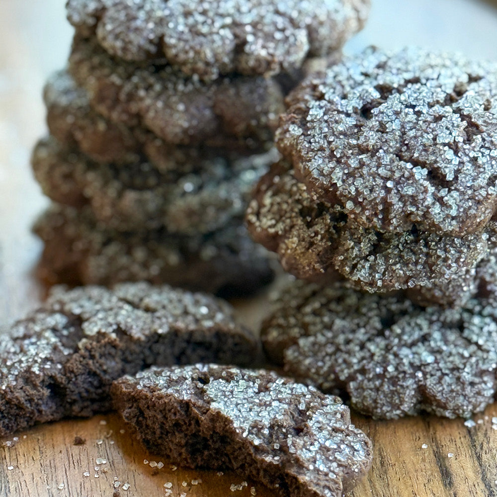 True Delicious Cocoa Blooms Cookies, topped with sparkling sugar, are stacked on a wooden surface; a few are broken open in front to reveal their soft, rich chocolate interior.