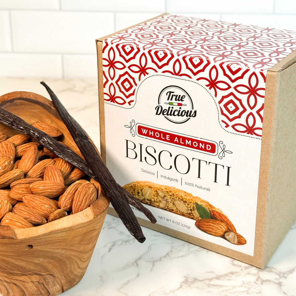 A box of True Delicious Elegant Box Almond Biscotti Individually Wrapped sits by whole almonds and vanilla beans on a marble countertop—an ideal Italian biscotti gift for any occasion.