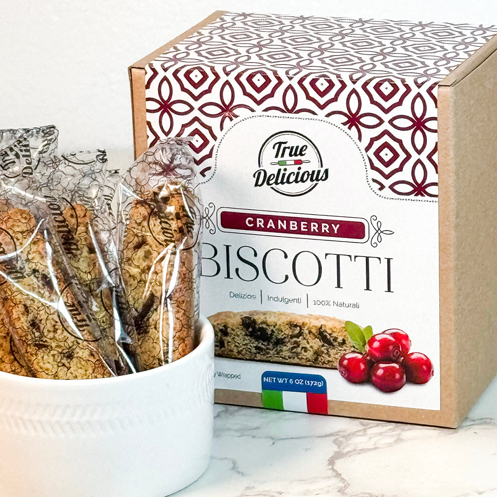 A box of True Delicious Elegant Box Cranberry Biscotti Individually Wrapped sits beside a white bowl with two wrapped treats. The box displays cranberry images, decorative patterns, and an Italian flag.