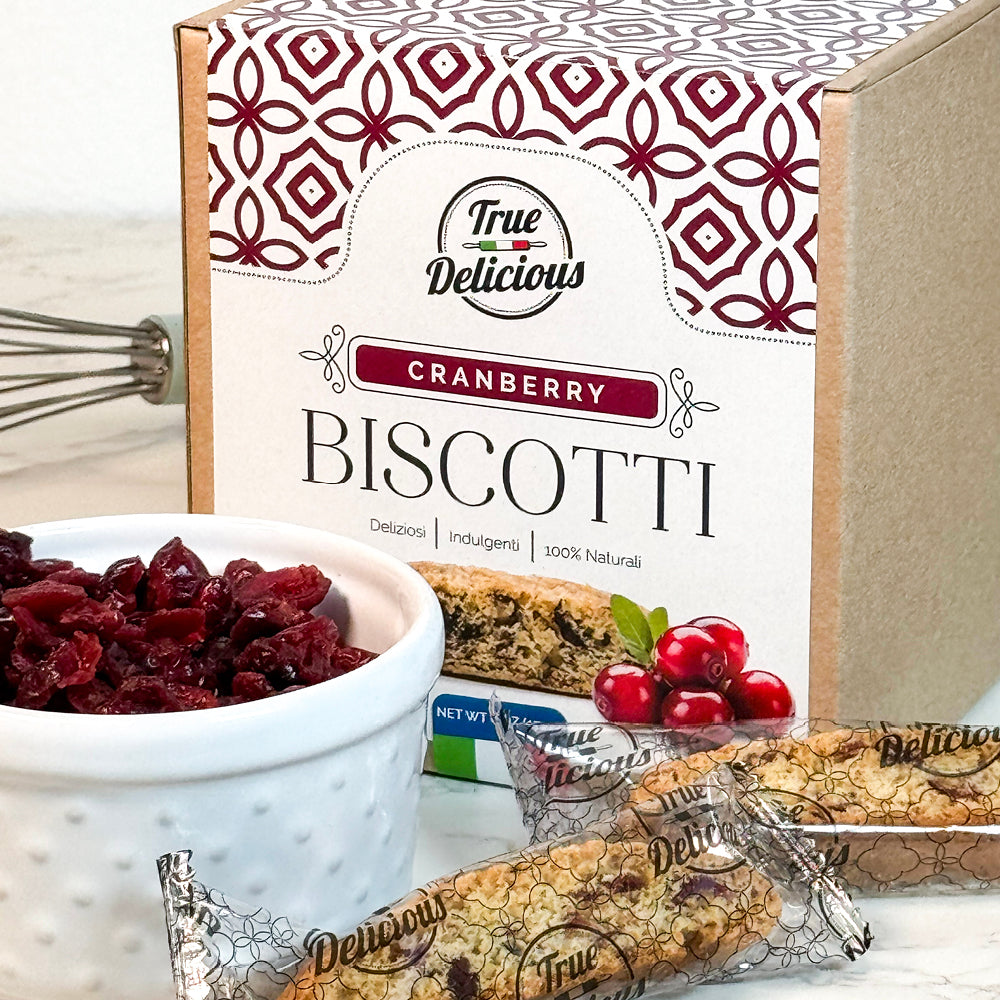 A box of True Delicious Elegant Box Cranberry Biscotti Individually Wrapped sits beside a white bowl of dried cranberries and two wrapped biscotti, with a whisk in the background.