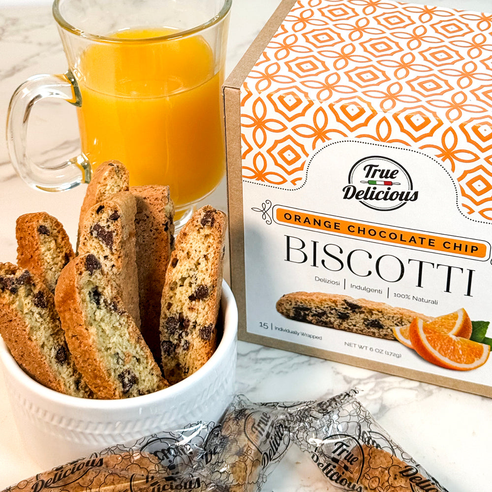 A cup of orange juice, a white bowl with artisan Italian biscotti, and an Elegant Box Orange Chocolate Chip Biscotti Individually Wrapped by True Delicious are displayed on a marble surface, with wrapped biscotti also visible.