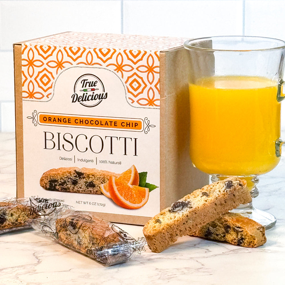 A box of True Delicious Elegant Box Orange Chocolate Chip Biscotti Individually Wrapped sits beside a glass of orange juice. Two Italian biscotti, one wrapped and one unwrapped, are displayed in front on a white surface.