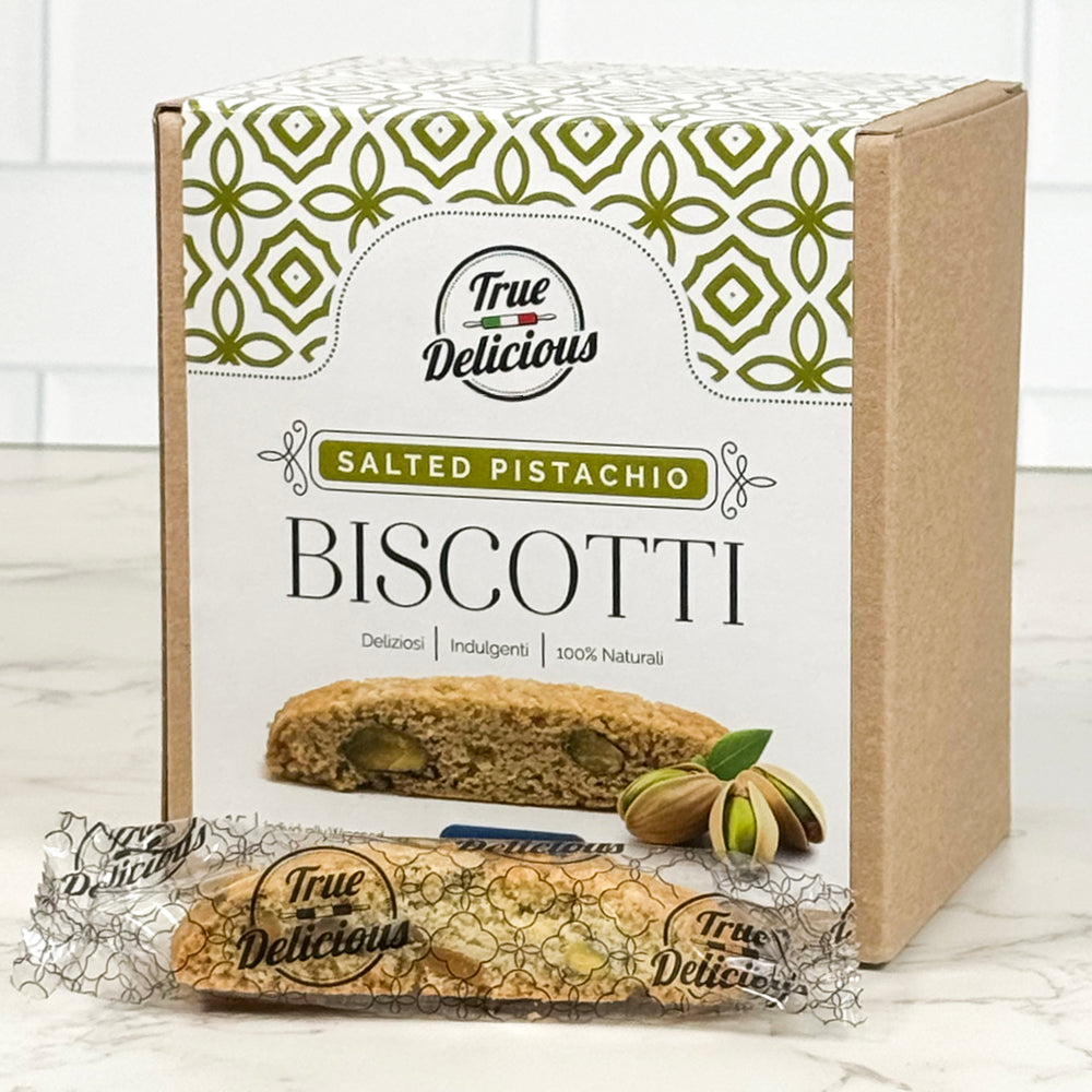A box of True Delicious Elegant Box Salted Pistachio Biscotti Individually Wrapped sits on the counter, with a single biscotto and pistachios in front. The packaging displays green patterns along with product details.