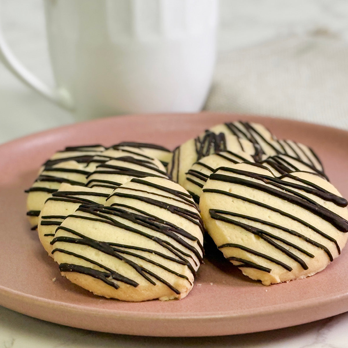 True Delicious Love Cookies, heart-shaped treats drizzled with chocolate, are arranged on a pink plate with a white mug in the background—perfect Valentine’s Day cookies for someone special.