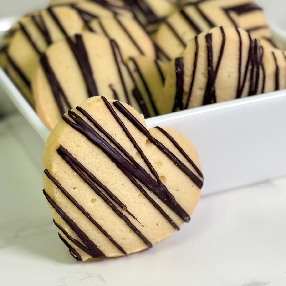 A heart-shaped Love Cookie by True Delicious, drizzled with dark chocolate, sits before a white dish filled with more of these Chocolate Drizzled Shortbread treats on a marble surface.