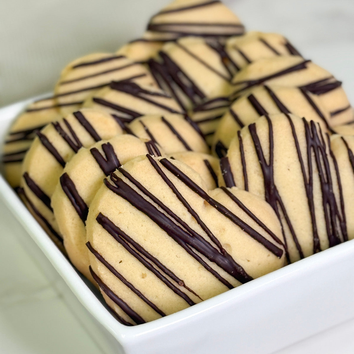 True Delicious Love Cookies: heart-shaped shortbread cookies with dark chocolate drizzle, neatly arranged in a white square bowl—perfect Valentine’s Day treats.